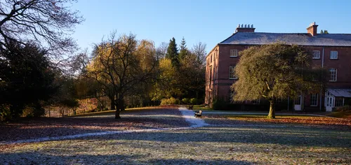 This landscape photograph captures Tapton House during a clear winter morning, with the grounds of Tapton Park in Chesterfield, United Kingdom visible in the foreground. Nature dominates the scene, with bare trees and scattered leaf litter across the frosty grass, highlighting the seasonal change. Tapton House, the main subject of the image, is a well-known building and landmark within the park, and stands prominently to the right, partially framed by mature trees. The early sunlight emphasizes the frost and shadows on the ground, enhancing the wintry atmosphere of the park. The presence of benches along the park path suggests a public space designed for visitors to enjoy the natural setting and historic architecture. The image represents a typical winter landscape found in British parks such as Tapton Park, blending trees, nature, and local history.